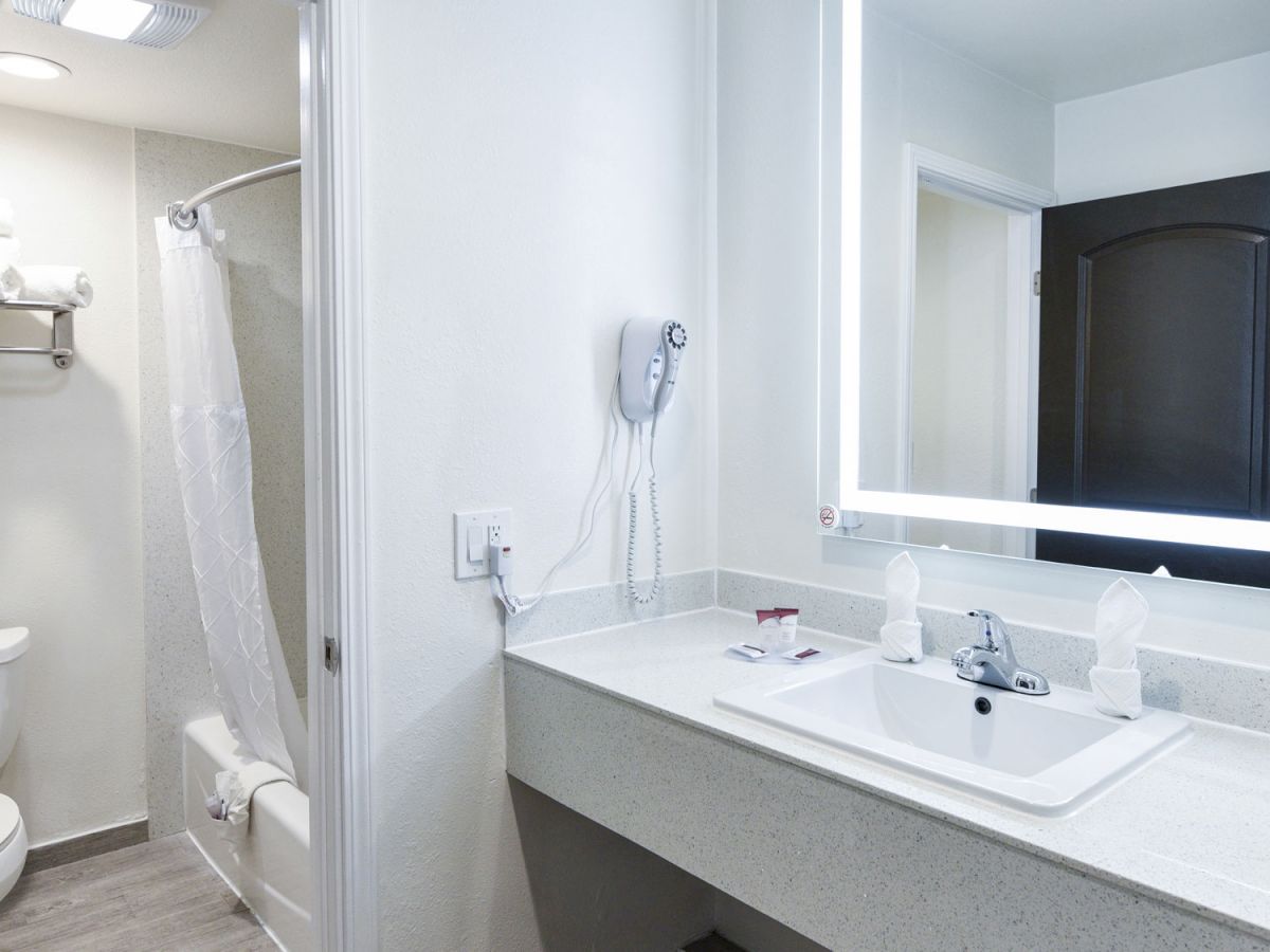 The image shows a modern bathroom with a sink, illuminated mirror, hairdryer, toilet, bathtub with shower curtain, and towels on a shelf.
