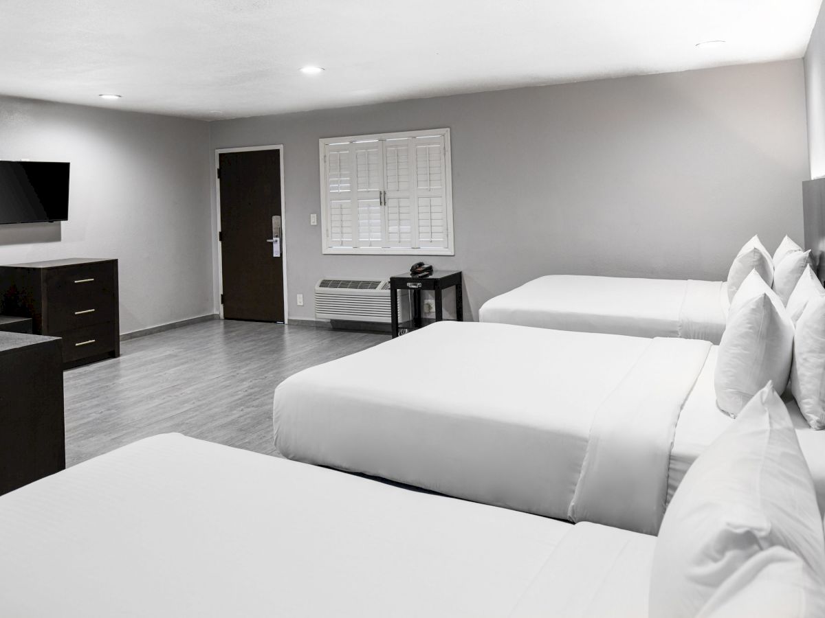 The image shows a hotel room with three double beds, a wall-mounted TV, a desk with a chair, and a wooden cabinet against a light grey wall.