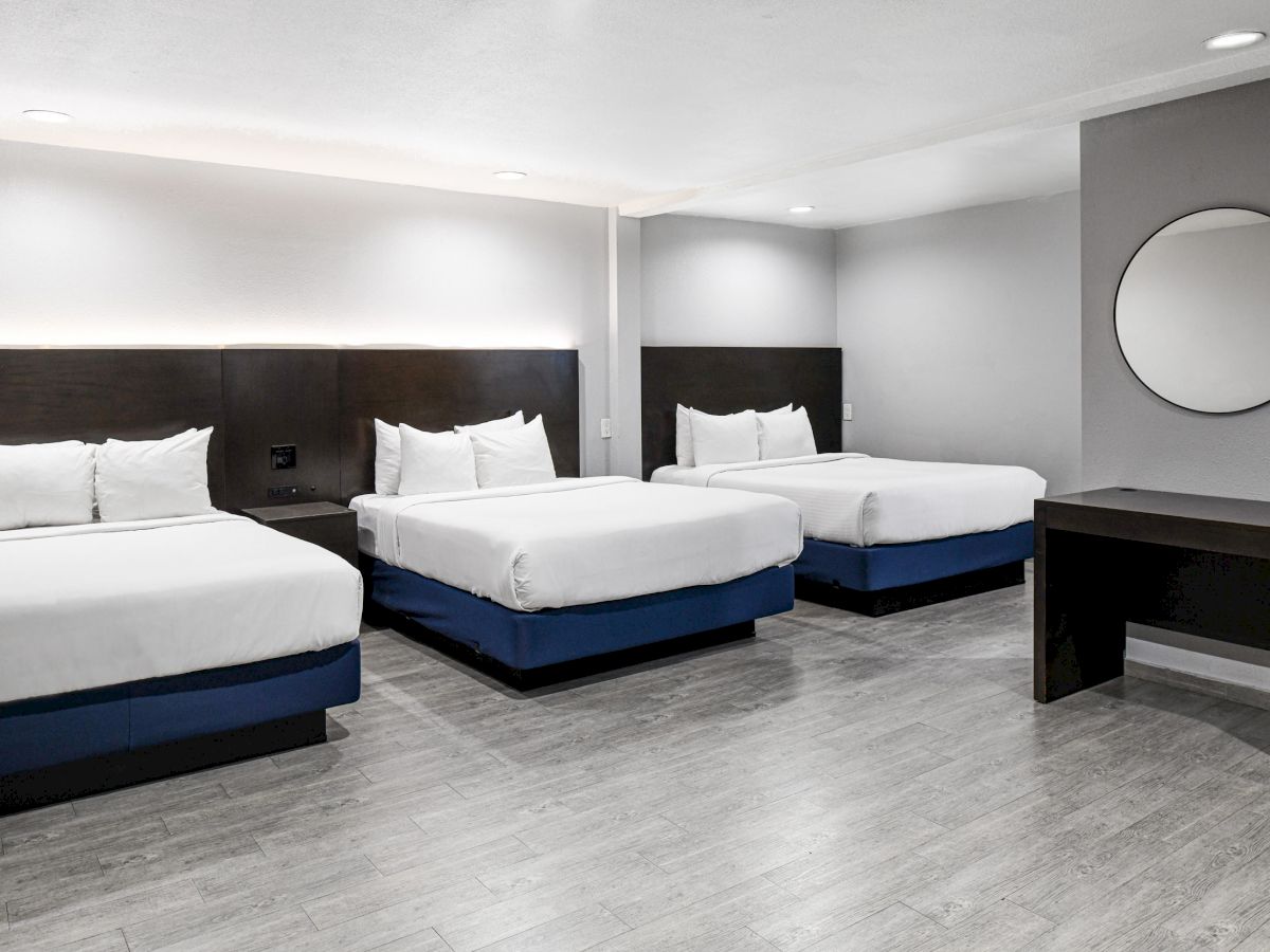 The image shows a modern hotel room with three neatly made double beds, gray walls, a large round mirror, and a dark wooden desk.