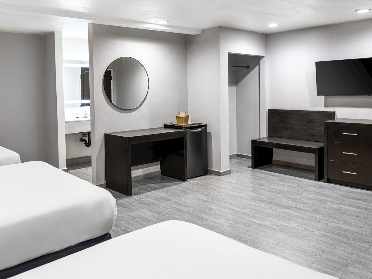 A clean, modern hotel room with three beds, a desk, a wall-mounted TV, a dresser, and a mirror. Gray tones dominate the color scheme.