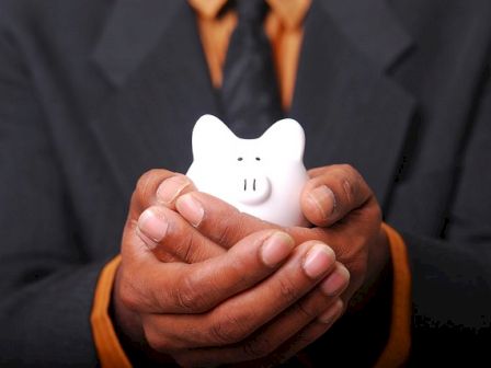 A person in a suit is holding a small white piggy bank with both hands, suggesting a concept of saving or financial security.