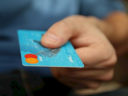 A hand is holding and extending a blue MasterCard credit card, symbolizing a financial transaction or payment being made.
