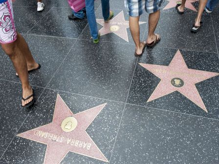 People walking on Hollywood Walk of Fame, with visible stars including Barbara Stanwyck and Drew Barrymore, on a tiled sidewalk.