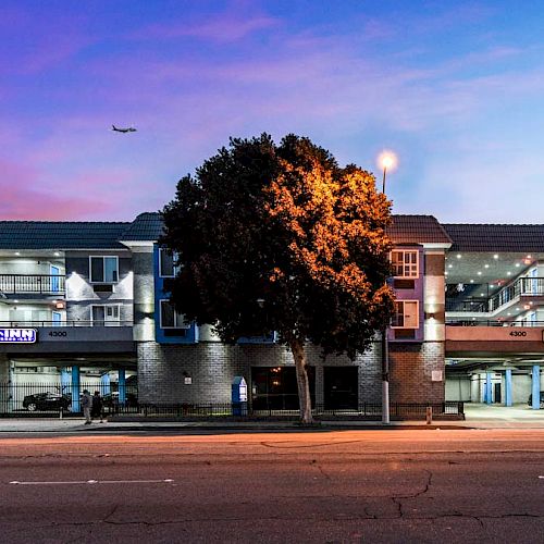 The image shows a three-story building with signage for an inn, illuminated at dusk with a tree on the sidewalk in front. An airplane is in the sky.
