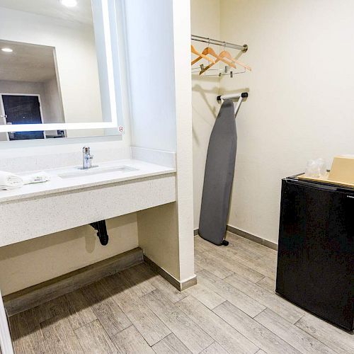 The image shows a small hotel room area with a sink, mirror, hair dryer, ironing board, hangers, mini fridge, and some disposable cups on the fridge.