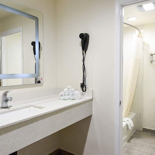 A clean, modern bathroom features a large mirror, hairdryer, toiletries on the counter, a sink, toilet, shower, and neatly folded towels on a shelf.