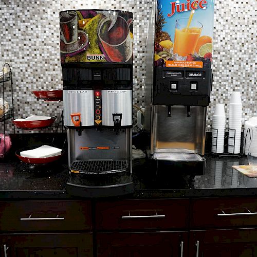 The image shows a self-serve beverage station with coffee and juice dispensers, paper cups, lids, a stack of plates, and a display of coffee creamers.