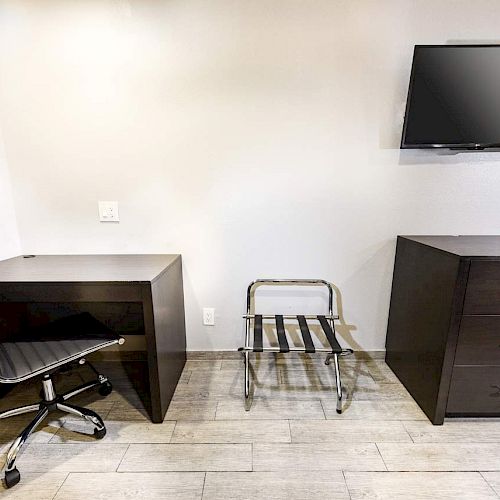The image shows a room with a desk and chair, a luggage stand, a chest of drawers, and a wall-mounted TV.