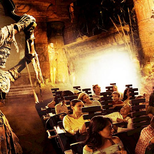 People seated on a theme park ride appear to be reacting to an animatronic skeleton warrior inside a dimly lit, adventurous setting.
