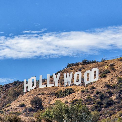 The image shows the iconic Hollywood sign situated on a hillside with greenery and a blue sky with some clouds in the background.