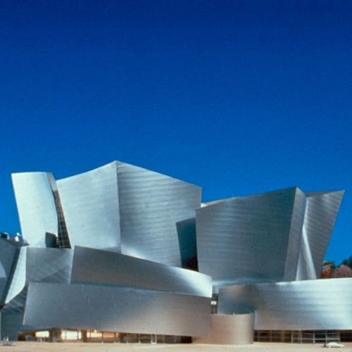 The image shows a modern, abstract architectural building with irregular shapes and metallic surfaces, set against a clear blue sky.
