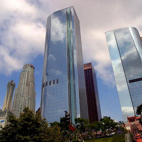 The image shows a skyline with several tall, reflective skyscrapers set against a partly cloudy sky with some trees and buildings in the foreground.