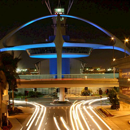 This image shows a night view of the iconic LAX Theme Building at Los Angeles International Airport, with light trails from vehicles on the road.