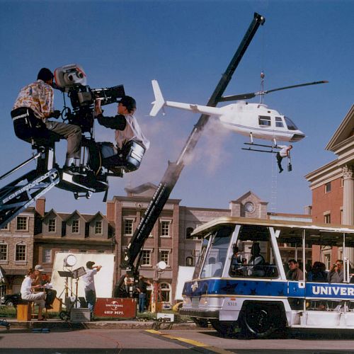 A film set with cameras, crew on cranes, a tram labeled "Universal Studios," and a helicopter suspended by a crane in front of a building.