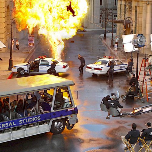 A group of tourists on a Universal Studios tram tour watches a staged explosion scene with police cars and film crew equipment nearby.