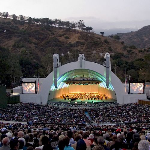 The image shows a large crowd seated in an outdoor amphitheater surrounded by hills, watching a performance on a stage with an iconic shell structure.