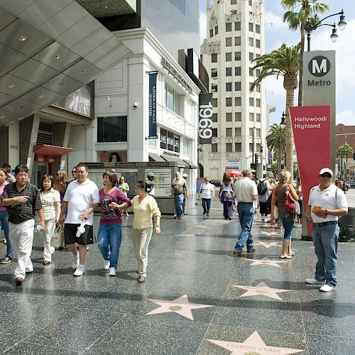 The image shows people walking on Hollywood Boulevard with the Hollywood Walk of Fame stars on the sidewalk and a Metro station sign in the background.