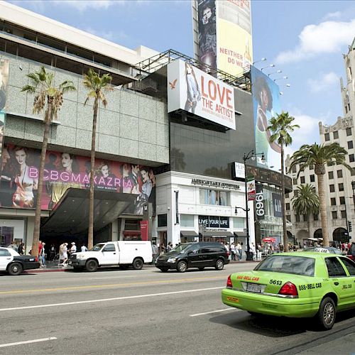 City street with various parked cars, including a green taxi. Buildings with large advertisements and palm trees, capturing an urban setting.