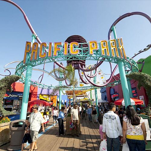 The image shows the entrance to Pacific Park on the Santa Monica Pier, featuring a large archway and Ferris wheel, with people walking around.