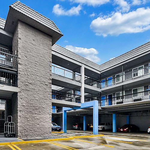 The image shows a parking area of a multi-story building with cars parked and staircases leading to upper floors under a partly cloudy sky.