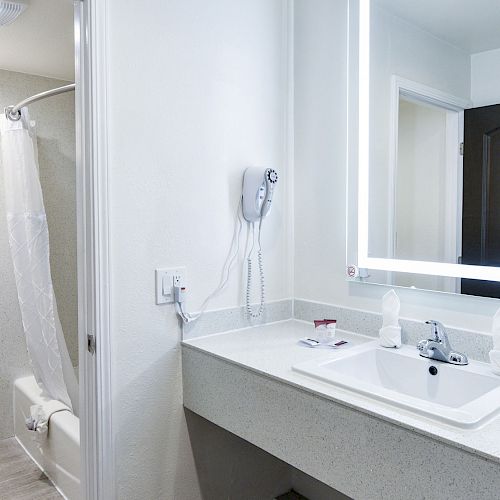 A modern bathroom with a sink, illuminated mirror, hairdryer, toilet, shower with curtain, and towel rack holding towels and toiletries.