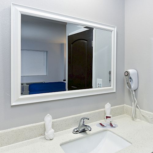 The image shows a bathroom sink with a mirror, a faucet, a hairdryer on the wall, and two rolled towels on the counter in a light-colored room.