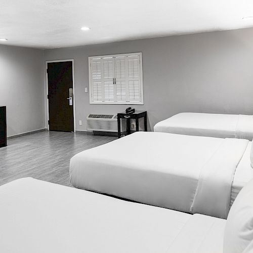 The image shows a clean, modern hotel room with three neatly made beds, a flat-screen TV mounted on the wall, a dresser, and a door in the background.