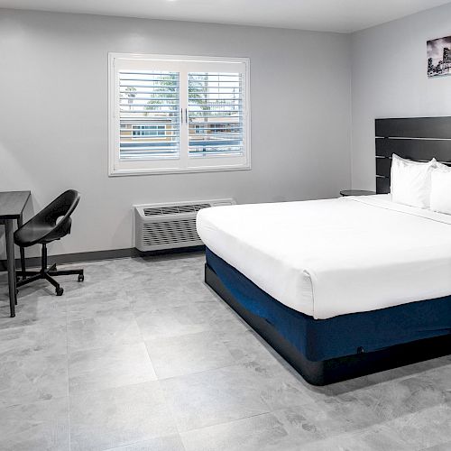 A modern, minimalist hotel room with a neatly made bed, desk, chair, and wall-mounted TV. The room has gray floors and a window with white blinds.