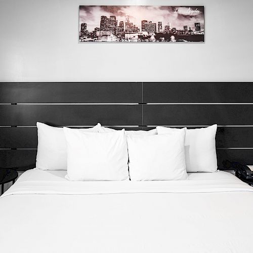 A neatly made bed with white linens and pillows, black headboard, side tables with objects, and a framed cityscape above the headboard.