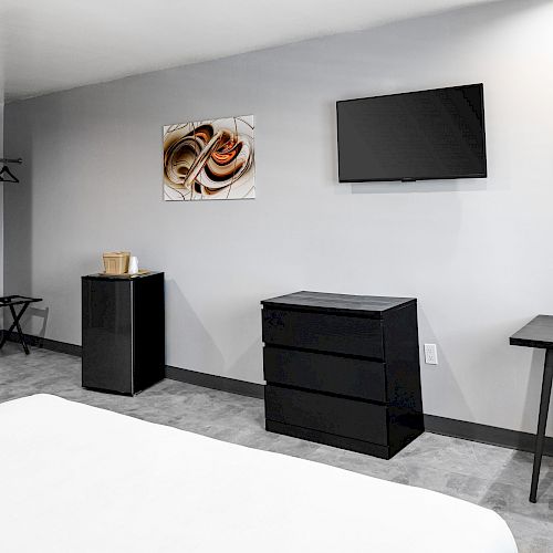 The image shows a modern hotel room with a TV, dresser, round mirror, desk, and wall art featuring cinnamon rolls.