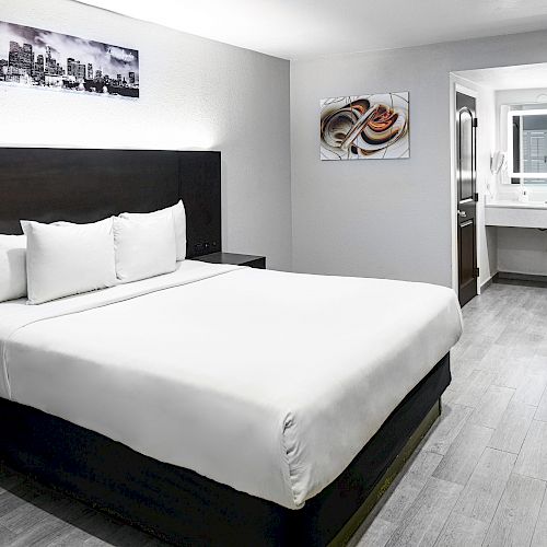 The image shows a modern hotel room with a large bed, artwork on the walls, a small work desk with a chair, and an en-suite bathroom in the background.