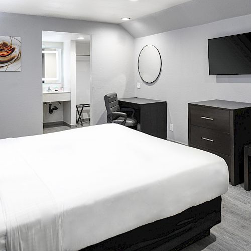 A modern hotel room has a bed, a wall-mounted TV, a desk with a chair, a dresser, a bench, a painting above the bed, and a bathroom in the background.