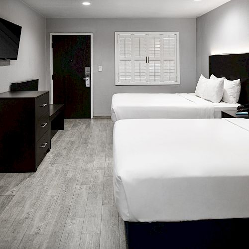 A modern hotel room features two neatly made beds, a wall-mounted TV, a dresser, and a door leading outside, all under ambient lighting.