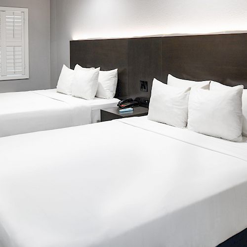 The image shows a modern hotel room with two neatly made, white-sheeted beds, a nightstand in between, and a window with closed shutters.