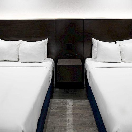 The image shows a modern hotel room with two neatly made double beds, white linens, and a shared nightstand with some decor in the middle.