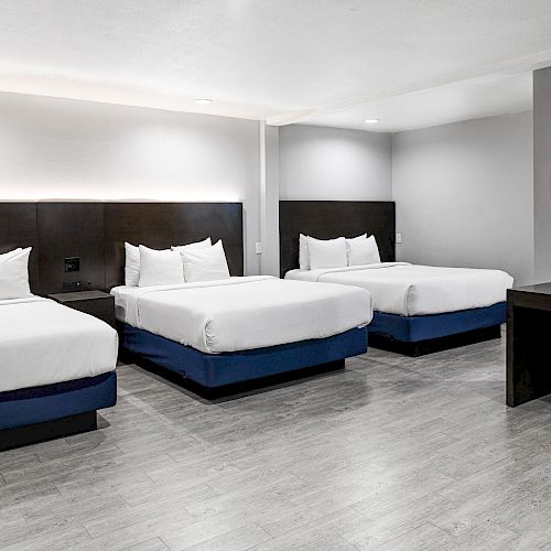 The image shows a modern hotel room with three queen-sized beds, a wooden floor, a desk with a mirror, and minimalistic decor.