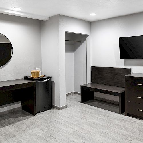 The image shows a modern room with a desk, a round mirror, a mini-fridge, a TV, a bench, and a dresser, all in black furniture.