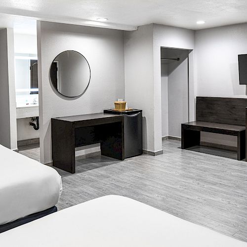 The image shows a modern hotel room with three beds, a desk with a round mirror, a TV mounted on the wall, and a bathroom visible in the background.