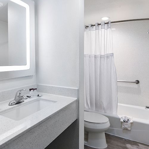 A clean, modern bathroom with a lit mirror, sink, and faucet, a toilet, a bathtub with a shower curtain, and a towel hanging on the bathtub's side.