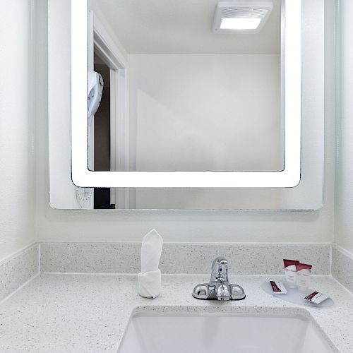 A modern bathroom countertop with a sink, illuminated mirror, wall-mounted hairdryer, tissue, tap, and toiletries laid on the right side ends the sentence.