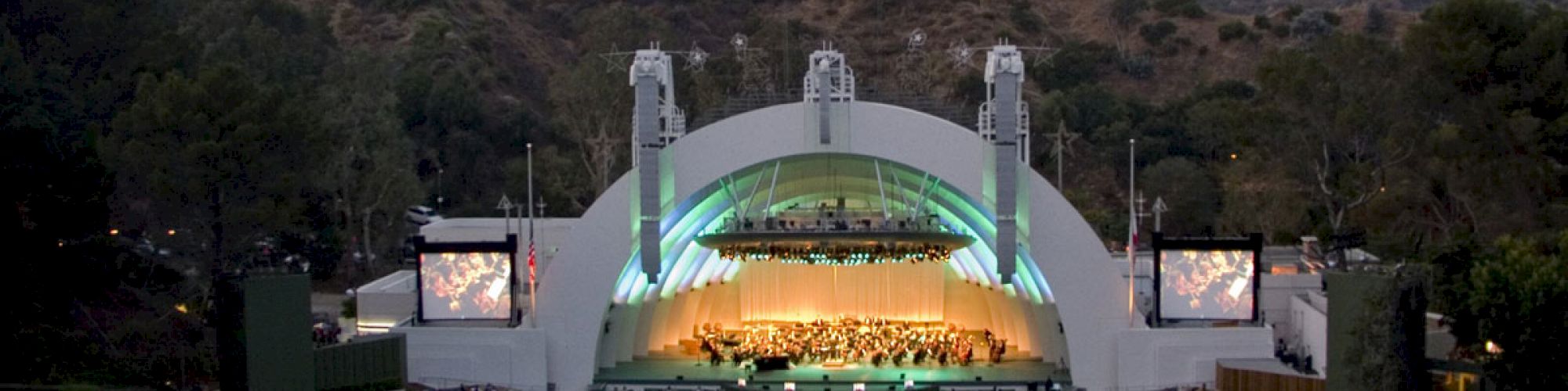 The image shows a large outdoor amphitheater amidst hills, with a seated audience watching a live performance on stage, under a lit dome structure.