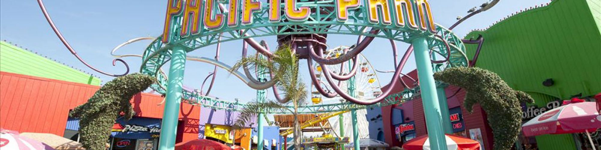 The image shows the entrance to Pacific Park, an amusement park with a roller coaster structure in view, located on a pier bustling with people.