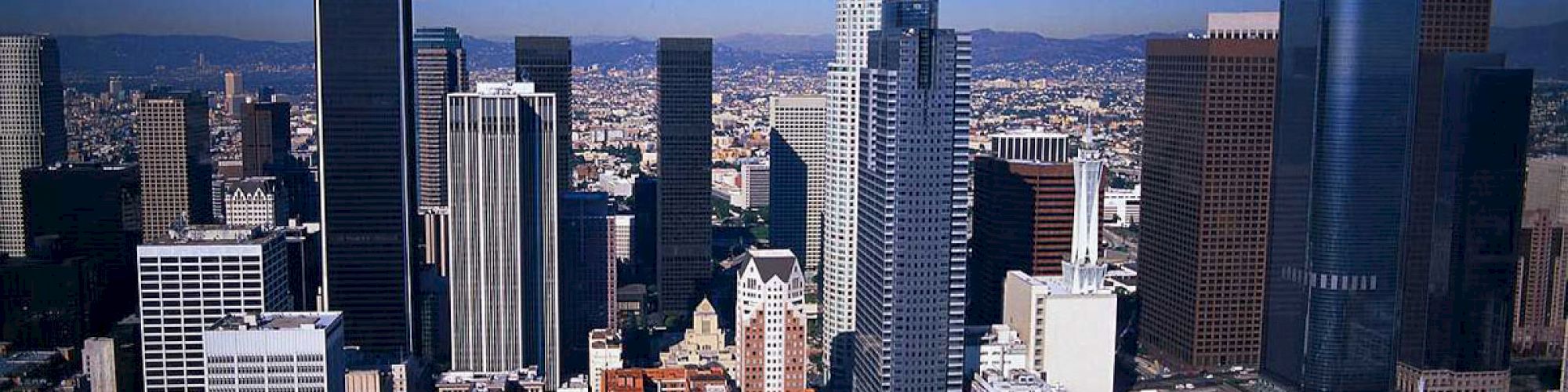 The image shows a cityscape with tall skyscrapers, including several modern glass buildings and older architecture, under a clear blue sky.