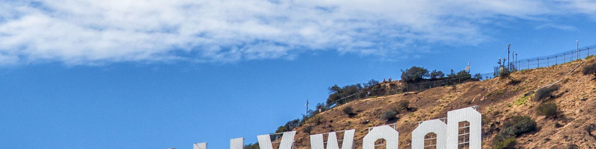 The image shows the famous Hollywood sign situated on a hillside with clear blue skies and some clouds in the background.