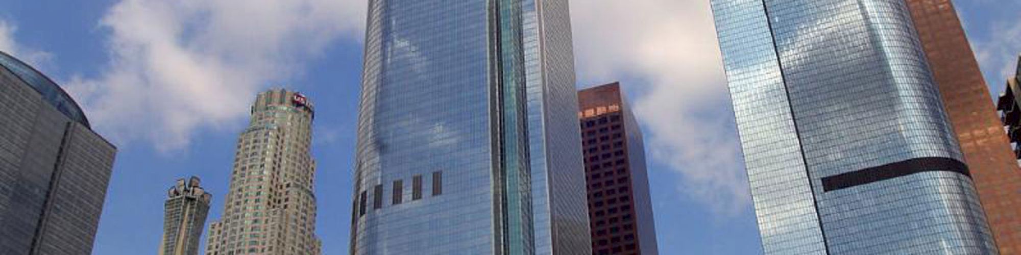 The image shows a cityscape with several modern skyscrapers and reflective glass buildings under a partly cloudy sky, surrounded by some trees.