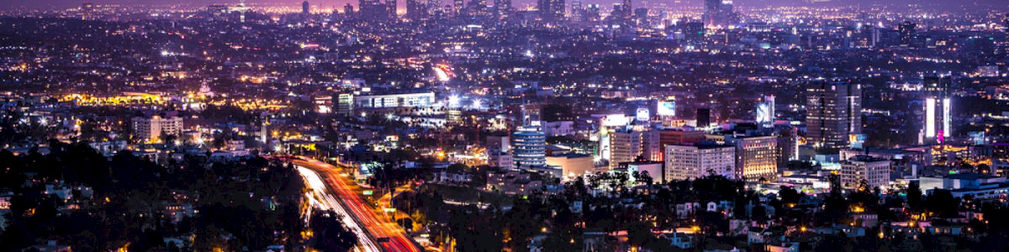 A nighttime cityscape featuring a skyline with tall buildings and a winding road lit by car lights, set against a deep blue and purple sky.