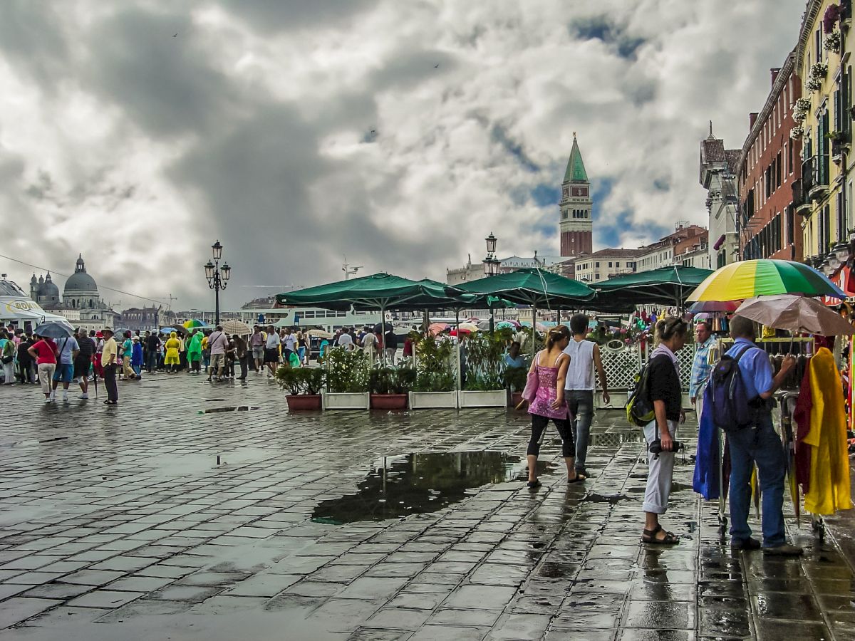 A busy open square with people walking, some with umbrellas. Green tents are set up, possibly for a market. Old buildings and a tower in the background.