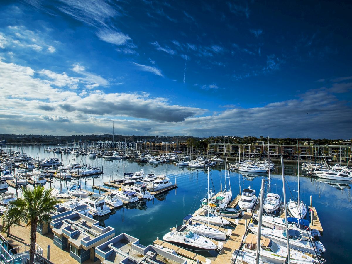 The image shows a marina filled with numerous yachts and sailboats docked at piers, under a partly cloudy blue sky reflecting on the water.