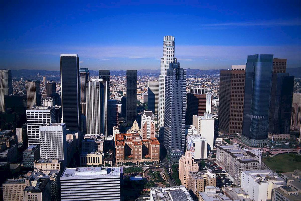 A city skyline with numerous tall buildings under a clear blue sky. The tallest building is uniquely designed, standing prominently among others.