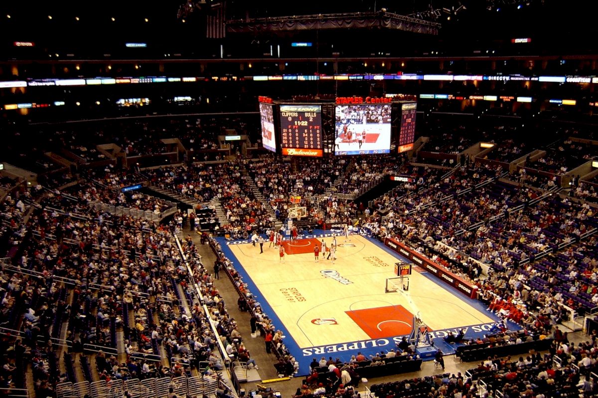 The image depicts an indoor basketball arena filled with spectators, featuring a game in progress and a large scoreboard displaying game information.